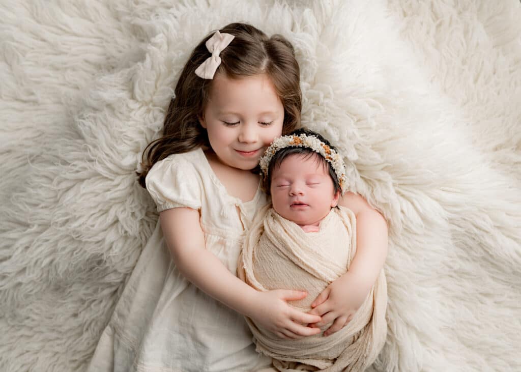 Older sister lying next to newborn baby, gently holding and smiling during a sibling photography session