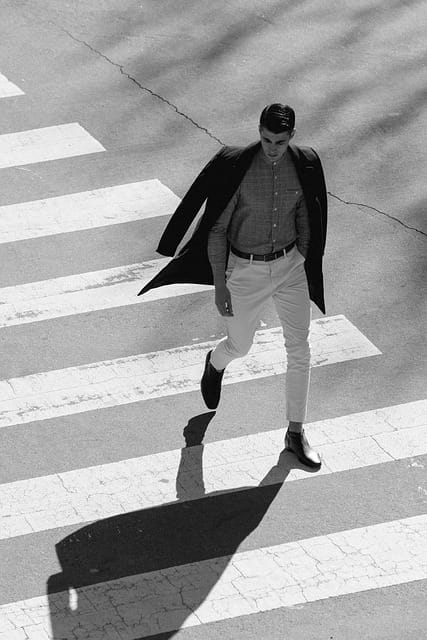 Man in a black coat walking on a crosswalk, wearing a checkered shirt and white trousers, ideal for stylish professional headshots male.