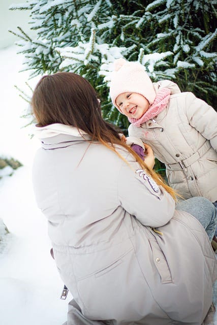 “Mother holding her baby in the snow, both bundled up in warm winter clothes, surrounded by a snowy landscape.”