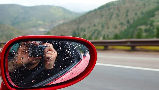 Close-up of a car’s side mirror with raindrops reflecting a person taking a photo, with scenic green mountains and highway in the background.