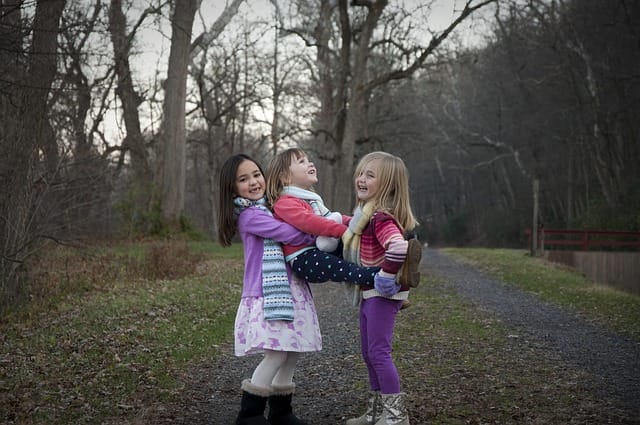 “Three little sisters posing outdoors in winter, wearing warm clothes, without scarves, standing on a clear ground with no snow around.